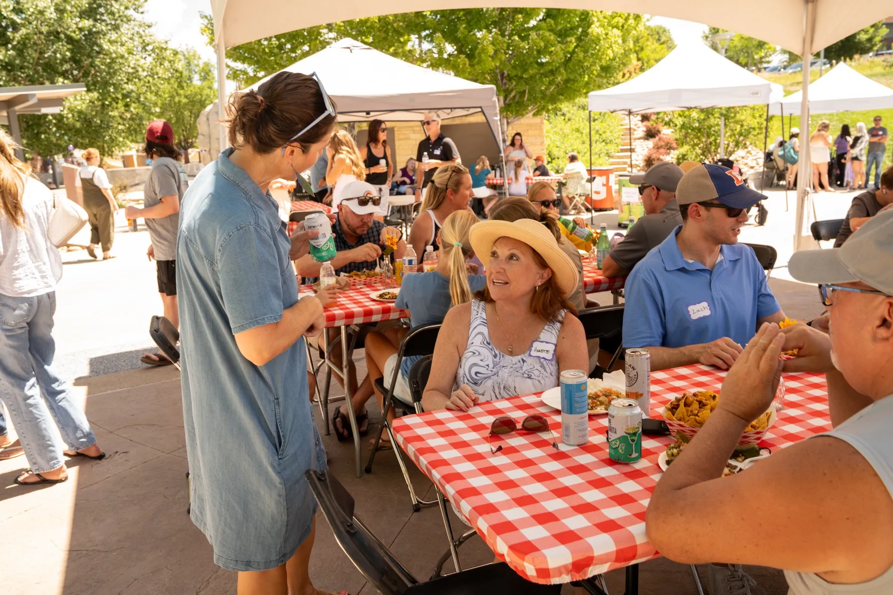 Team members sharing a meal at red-checked picnic tables under shade tents